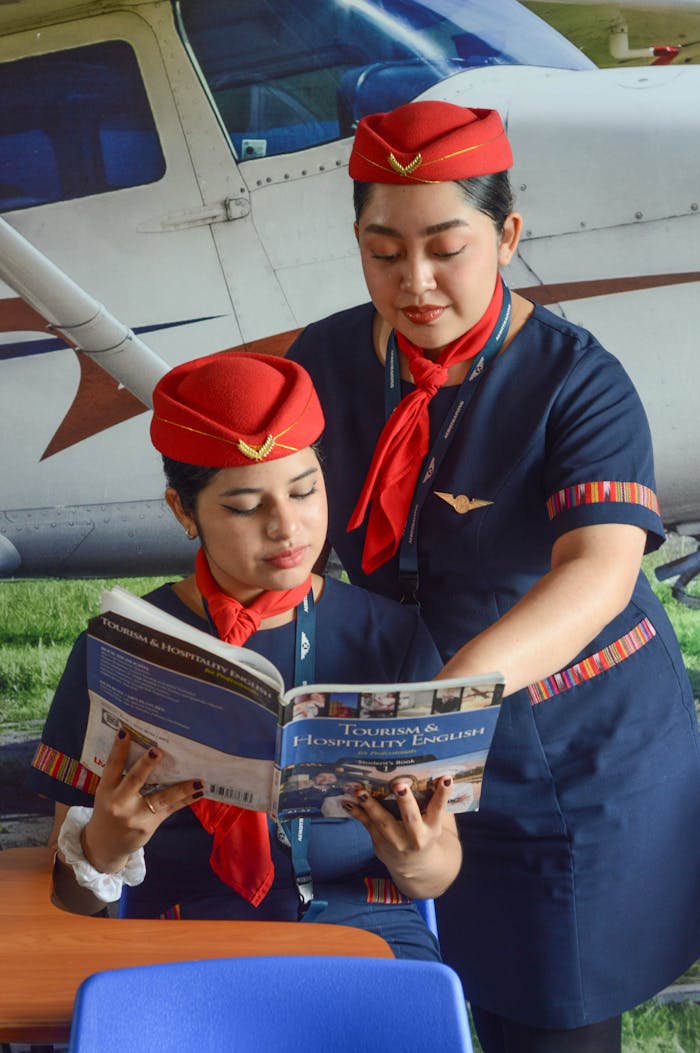 who-we-are Two flight attendants in uniform reading aviation-related books outdoors near a plane.